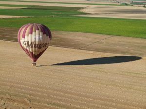 Un globo aerostático con Muga a bordo se desplaza suavemente sobre un vasto campo en La Rioja, proyectando una larga sombra que recuerda a la aventura Vuelo en globo por La Rioja con Globos Arcoiris. - ForTwo - Planes para parejas