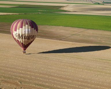 Un globo aerostático con Muga a bordo se desplaza suavemente sobre un vasto campo en La Rioja, proyectando una larga sombra que recuerda a la aventura Vuelo en globo por La Rioja con Globos Arcoiris. - ForTwo - Planes para parejas