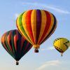 Tres globos aerostáticos con una paleta de colores vibrantes ascienden con gracia hacia el cielo azul claro, brindando una vista impresionante de la Sierra de Guadarrama durante su "Vuelo en globo por el paisaje madrileño y la Sierra de Guadarrama". - ForTwo - Planes para parejas