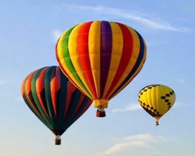 Tres globos aerostáticos con una paleta de colores vibrantes ascienden con gracia hacia el cielo azul claro, brindando una vista impresionante de la Sierra de Guadarrama durante su "Vuelo en globo por el paisaje madrileño y la Sierra de Guadarrama". - ForTwo - Planes para parejas