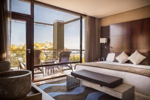 Descubra la elegancia en la Gran Suite Mediterránea con terraza en el hotel Miramar (Barcelona), una moderna habitación con una gran cama, almohadas decorativas y una terraza con vistas a frondosos árboles y aguas serenas. - ForTwo - Planes para parejas