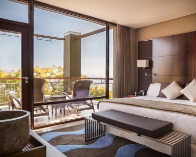 Descubra la elegancia en la Gran Suite Mediterránea con terraza en el hotel Miramar (Barcelona), una moderna habitación con una gran cama, almohadas decorativas y una terraza con vistas a frondosos árboles y aguas serenas. - ForTwo - Planes para parejas