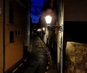 Un callejón estrecho al anochecer, iluminado por farolas que proyectan un brillo misterioso sobre los edificios, crea el escenario perfecto para el Tour nocturno por la Granada Paranormal contra un cielo azul oscuro. - ForTwo - Planes para parejas