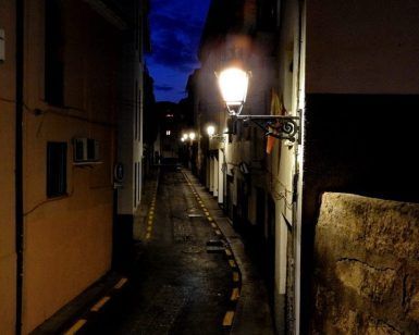 Un callejón estrecho al anochecer, iluminado por farolas que proyectan un brillo misterioso sobre los edificios, crea el escenario perfecto para el Tour nocturno por la Granada Paranormal contra un cielo azul oscuro. - ForTwo - Planes para parejas