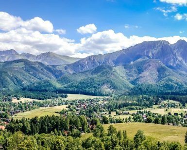 La "Visita guiada a Zakopane y a las Tatras (Cracovia)" muestra un paisaje montañoso con exuberante vegetación, un pintoresco pueblo y un cielo azul salpicado de nubes. - ForTwo - Planes para parejas