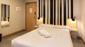 Disfruta de una moderna habitación de hotel en Bilbao con una cama impecablemente hecha, tres toallas enrolladas, un cabecero a rayas y una puerta a la izquierda. Ideal para viajeros que buscan comodidad en el Hostel Bcool Bilbao. - ForTwo - Planes para parejas