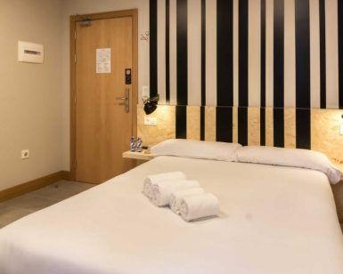 Disfruta de una moderna habitación de hotel en Bilbao con una cama impecablemente hecha, tres toallas enrolladas, un cabecero a rayas y una puerta a la izquierda. Ideal para viajeros que buscan comodidad en el Hostel Bcool Bilbao. - ForTwo - Planes para parejas