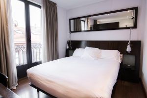 Disfrute de una estancia de ensueño en el Hotel Petit Palace, Bilbao. Esta elegante habitación incluye una cómoda cama doble con sábanas blancas, un cabecero oscuro y un gran espejo. Las ventanas con cortinas realzan la sensación de amplitud, complementando a la perfección el cálido suelo de madera. - ForTwo - Planes para parejas