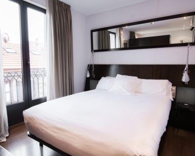 Disfrute de una estancia de ensueño en el Hotel Petit Palace, Bilbao. Esta elegante habitación incluye una cómoda cama doble con sábanas blancas, un cabecero oscuro y un gran espejo. Las ventanas con cortinas realzan la sensación de amplitud, complementando a la perfección el cálido suelo de madera. - ForTwo - Planes para parejas