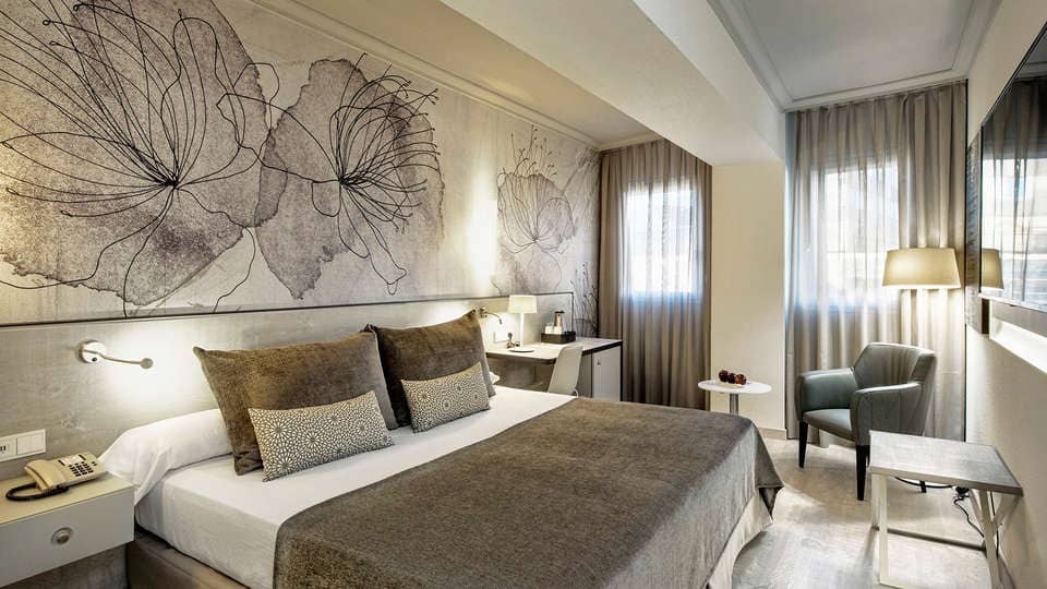 Disfrute de la escapada perfecta en una habitación moderna del Hotel Escapada Sallés Pere IV de Barcelona, con una cama grande, arte floral, un cómodo sillón, lámpara y ventanas con cortinas transparentes que dejan entrar la luz natural. El paquete incluye desayuno, acceso al spa y bebida de bienvenida. - ForTwo - Planes para parejas