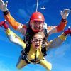 Dos paracaidistas en tándem en el cielo azul claro, con cascos y monos y un avión encima: una experiencia emocionante con Salto en paracaídas con Skydive Madrid, flotando elegantemente hacia la tierra. - ForTwo - Planes para parejas