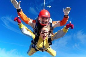 Dos paracaidistas en tándem en el cielo azul claro, con cascos y monos y un avión encima: una experiencia emocionante con Salto en paracaídas con Skydive Madrid, flotando elegantemente hacia la tierra. - ForTwo - Planes para parejas