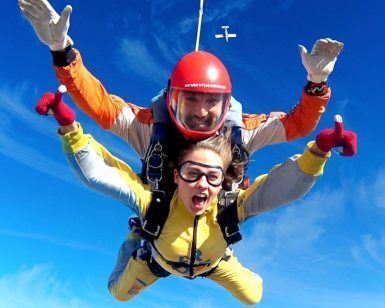 Dos paracaidistas en tándem en el cielo azul claro, con cascos y monos y un avión encima: una experiencia emocionante con Salto en paracaídas con Skydive Madrid, flotando elegantemente hacia la tierra. - ForTwo - Planes para parejas