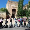 Los segways se deslizan con gracia junto a un antiguo edificio de piedra con una entrada arqueada en medio de la exuberante vegetación del Realejo, evocando el encanto atemporal de la Alhambra. Vívelo con "Hacer una ruta en Segway por la Alhambra y el Realejo". - ForTwo - Planes para parejas