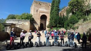 Los segways se deslizan con gracia junto a un antiguo edificio de piedra con una entrada arqueada en medio de la exuberante vegetación del Realejo, evocando el encanto atemporal de la Alhambra. Vívelo con "Hacer una ruta en Segway por la Alhambra y el Realejo". - ForTwo - Planes para parejas