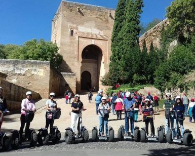 Los segways se deslizan con gracia junto a un antiguo edificio de piedra con una entrada arqueada en medio de la exuberante vegetación del Realejo, evocando el encanto atemporal de la Alhambra. Vívelo con "Hacer una ruta en Segway por la Alhambra y el Realejo". - ForTwo - Planes para parejas