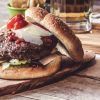 Deléitate con una jugosa hamburguesa gourmet con lechuga, queso y tomate sobre una tabla de madera, acompañada de papas fritas y cerveza fría. ¡Experimenta el "Curso de preparación de hamburguesas premium" en Madrid para quienes buscan sabores gourmet!. - ForTwo - Planes para parejas