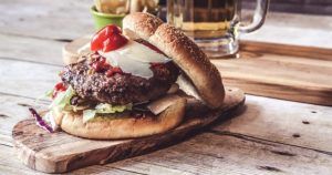 Deléitate con una jugosa hamburguesa gourmet con lechuga, queso y tomate sobre una tabla de madera, acompañada de papas fritas y cerveza fría. ¡Experimenta el "Curso de preparación de hamburguesas premium" en Madrid para quienes buscan sabores gourmet!. - ForTwo - Planes para parejas