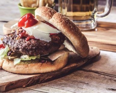 Deléitate con una jugosa hamburguesa gourmet con lechuga, queso y tomate sobre una tabla de madera, acompañada de papas fritas y cerveza fría. ¡Experimenta el "Curso de preparación de hamburguesas premium" en Madrid para quienes buscan sabores gourmet!. - ForTwo - Planes para parejas