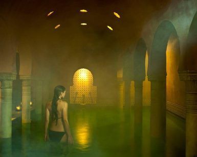 Una persona en traje de baño se encuentra en una piscina cubierta tenue y brumosa en Hammam Al Ándalus Granada Baños Árabes, rodeada de arcos y luces suaves y brillantes. - ForTwo - Planes para parejas