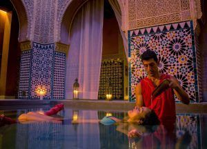 Un hombre vierte suavemente agua sobre la cabeza de una mujer mientras ella flota pacíficamente en una lujosa y ornamentada piscina cubierta, evocando la relajante experiencia del Baño con masaje MIMMA 15 en Hammam Al Ándalus Málaga. - ForTwo - Planes para parejas