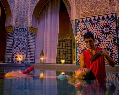 Un hombre vierte suavemente agua sobre la cabeza de una mujer mientras ella flota pacíficamente en una lujosa y ornamentada piscina cubierta, evocando la relajante experiencia del Baño con masaje MIMMA 15 en Hammam Al Ándalus Málaga. - ForTwo - Planes para parejas
