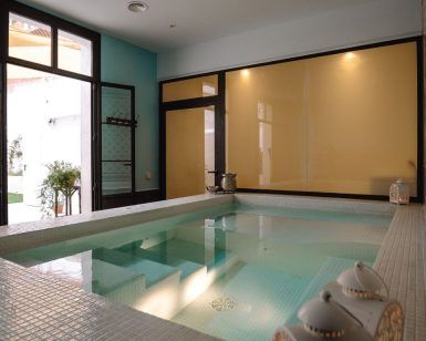 La sala de la piscina cubierta cuenta con paredes de azulejos, detalles en verde azulado y amplios ventanales. Las teteras junto a la piscina transmiten una atmósfera acogedora que evoca una tarde tranquila en "Mil y una noches a dúo - Hammam en Le Patio Spa". - ForTwo - Planes para parejas