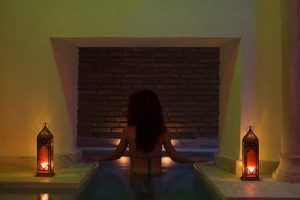 Alguien se relaja en una piscina de hidromasaje suavemente iluminada en Hammam con masaje de 15 minutos en los baños árabes de Al-Haram (Granada), donde las velas parpadeantes proyectan suaves sombras sobre la pared de ladrillos, evocando los tradicionales baños árabes. - ForTwo - Planes para parejas