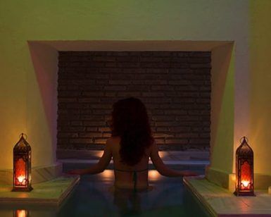 Alguien se relaja en una piscina de hidromasaje suavemente iluminada en Hammam con masaje de 15 minutos en los baños árabes de Al-Haram (Granada), donde las velas parpadeantes proyectan suaves sombras sobre la pared de ladrillos, evocando los tradicionales baños árabes. - ForTwo - Planes para parejas