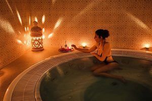 Una mujer en bikini se relaja en un jacuzzi, rodeada de velas y faroles ornamentados, creando la encantadora atmósfera de un Baño árabe en pareja con masaje sencillo en Medina Aljarafe (Sevilla). - ForTwo - Planes para parejas