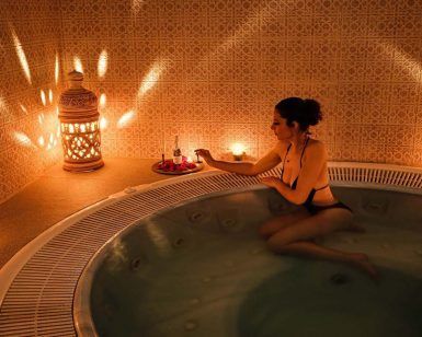 Una mujer en bikini se relaja en un jacuzzi, rodeada de velas y faroles ornamentados, creando la encantadora atmósfera de un Baño árabe en pareja con masaje sencillo en Medina Aljarafe (Sevilla). - ForTwo - Planes para parejas