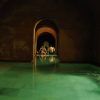 Una piscina cubierta tenuemente iluminada, con arcos en las puertas y aguas verdes, evoca la tranquilidad de un baño árabe. Al fondo, la gente se relaja, disfrutando de la serenidad como en la "Oferta Baño árabe con masaje en Hammam Al Ándalus Madrid". - ForTwo - Planes para parejas