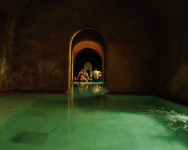 Una piscina cubierta tenuemente iluminada, con arcos en las puertas y aguas verdes, evoca la tranquilidad de un baño árabe. Al fondo, la gente se relaja, disfrutando de la serenidad como en la "Oferta Baño árabe con masaje en Hammam Al Ándalus Madrid". - ForTwo - Planes para parejas