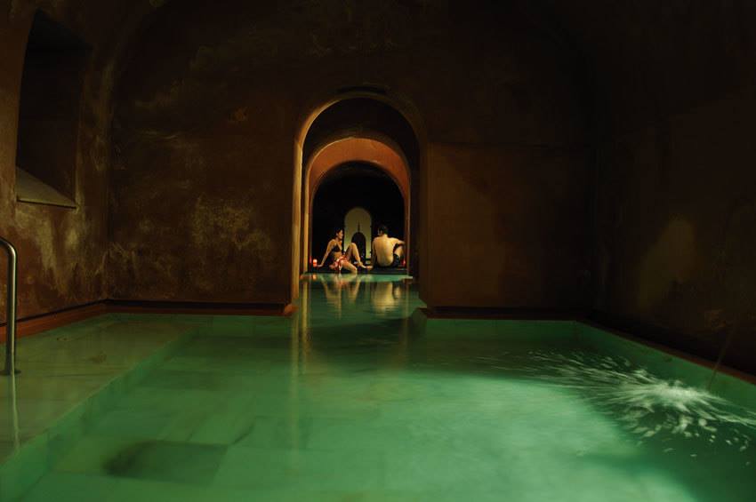 Una piscina cubierta tenuemente iluminada, con arcos en las puertas y aguas verdes, evoca la tranquilidad de un baño árabe. Al fondo, la gente se relaja, disfrutando de la serenidad como en la "Oferta Baño árabe con masaje en Hammam Al Ándalus Madrid". - ForTwo - Planes para parejas