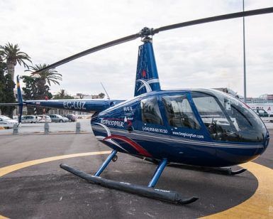 Un elegante helicóptero azul en el helipuerto, enmarcado por palmeras y un barco distante, inspira sueños de un emocionante "Vuelo en helicóptero sobre el Cañón Tajo de los Vados", que ofrece impresionantes vistas. - ForTwo - Planes para parejas