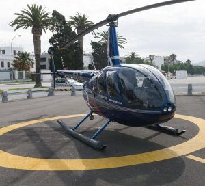 Un "Vuelo en helicóptero sobre el sur de Granada" en azul descansa sobre un helipuerto iluminado por el sol, envuelto por palmeras ondulantes y edificios de la ciudad, capturando la esencia vibrante de la región. - ForTwo - Planes para parejas