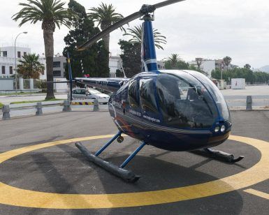 Un "Vuelo en helicóptero sobre el sur de Granada" en azul descansa sobre un helipuerto iluminado por el sol, envuelto por palmeras ondulantes y edificios de la ciudad, capturando la esencia vibrante de la región. - ForTwo - Planes para parejas