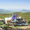 Bajo un cielo azul claro, la estructura metálica y ondulada de la Bodega Marqués de Riscal en Álava se alza entre viñedos, invitándote a una inolvidable Visita a la bodega Marqués de Riscal (Álava). - ForTwo - Planes para parejas