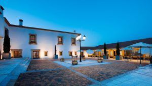 El patio de Escapada Hospes Palacio de Arenales, con sus paredes blancas y sus brillantes farolas, ofrece mesas cubiertas por sombrillas que evocan el ambiente sereno de un encantador desayuno. - ForTwo - Planes para parejas