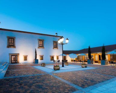 El patio de Escapada Hospes Palacio de Arenales, con sus paredes blancas y sus brillantes farolas, ofrece mesas cubiertas por sombrillas que evocan el ambiente sereno de un encantador desayuno. - ForTwo - Planes para parejas