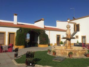El patio del Hotel El Moral exhibe una encantadora fuente de piedra, rodeada de mesas, botellas de vino y un edificio blanco y fresco bajo el cielo azul, ideal para la "Escapada romántica al hotel El Moral con visita a sus bodegas y cata de vinos (Badajoz). - ForTwo - Planes para parejas