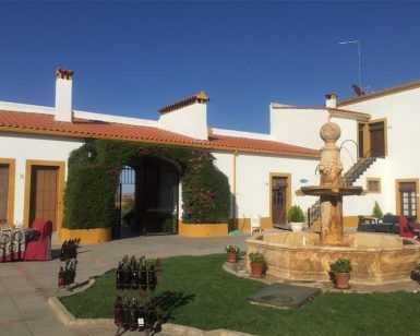 El patio del Hotel El Moral exhibe una encantadora fuente de piedra, rodeada de mesas, botellas de vino y un edificio blanco y fresco bajo el cielo azul, ideal para la "Escapada romántica al hotel El Moral con visita a sus bodegas y cata de vinos (Badajoz). - ForTwo - Planes para parejas
