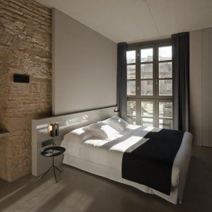 El dormitorio minimalista del Hotel Caro en Valencia incluye una cama grande, una pared con acento de ladrillo y cortinas en una ventana que proyectan sombras intrigantes en el suelo. - ForTwo - Planes para parejas