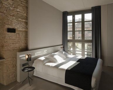 El dormitorio minimalista del Hotel Caro en Valencia incluye una cama grande, una pared con acento de ladrillo y cortinas en una ventana que proyectan sombras intrigantes en el suelo. - ForTwo - Planes para parejas