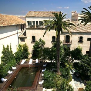 En el Hospes Palacio del Bailío (Córdoba), disfrute de un fin de semana revitalizante en un tranquilo patio con piscina, exuberante vegetación y edificios históricos. Las tumbonas le invitan a relajarse bajo el cielo azul despejado. - ForTwo - Planes para parejas