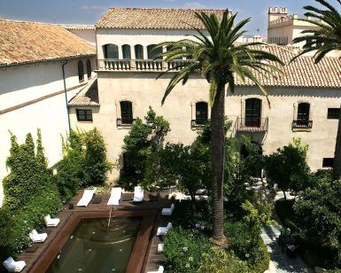 En el Hospes Palacio del Bailío (Córdoba), disfrute de un fin de semana revitalizante en un tranquilo patio con piscina, exuberante vegetación y edificios históricos. Las tumbonas le invitan a relajarse bajo el cielo azul despejado. - ForTwo - Planes para parejas