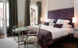 El elegante dormitorio, con cama doble, detalles en morado y un ambiente de spa, ofrece un "Fin de semana de lujo con spa en el Hotel Hospes Puerta Alcalá (Madrid)". Dos sillas y una mesa de cristal realzan los amplios ventanales con cortinas, ideales para relajarse. - ForTwo - Planes para parejas