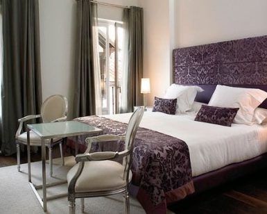 El elegante dormitorio, con cama doble, detalles en morado y un ambiente de spa, ofrece un "Fin de semana de lujo con spa en el Hotel Hospes Puerta Alcalá (Madrid)". Dos sillas y una mesa de cristal realzan los amplios ventanales con cortinas, ideales para relajarse. - ForTwo - Planes para parejas