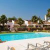 Una pintoresca escena de piscina al aire libre en Sotogrande cuenta con tumbonas y una persona descansando entre palmeras, bajo un cielo despejado y en medio de majestuosos edificios blancos: una escapada de fin de semana perfecta con una elegante cena bajo las estrellas, complementada con una botella de cava. - ForTwo - Planes para parejas