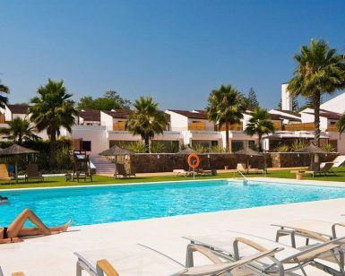 Una pintoresca escena de piscina al aire libre en Sotogrande cuenta con tumbonas y una persona descansando entre palmeras, bajo un cielo despejado y en medio de majestuosos edificios blancos: una escapada de fin de semana perfecta con una elegante cena bajo las estrellas, complementada con una botella de cava. - ForTwo - Planes para parejas